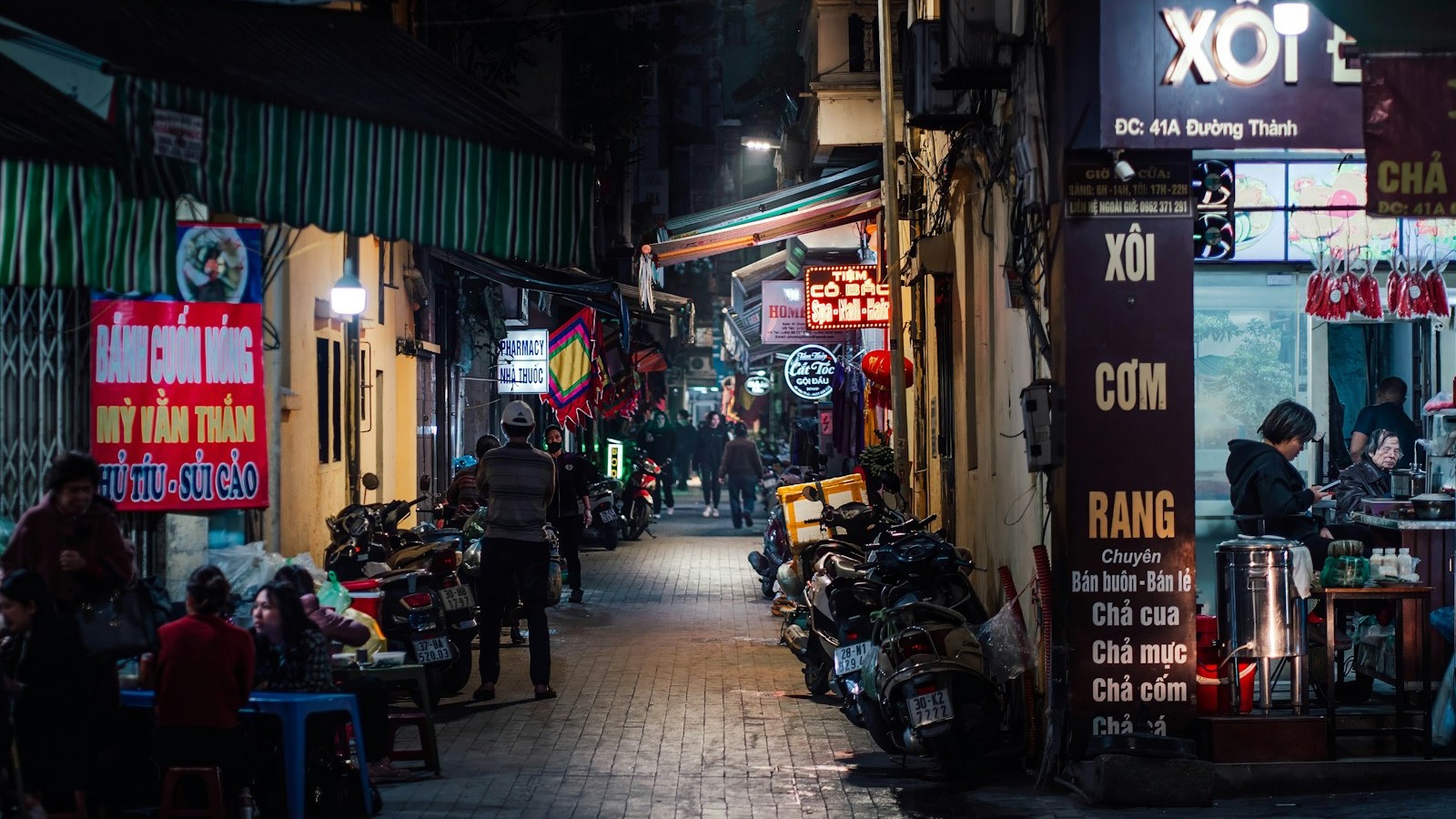 A narrow Vietnamese street at night, lit by shop signs and street lamps, with people seated at low tables